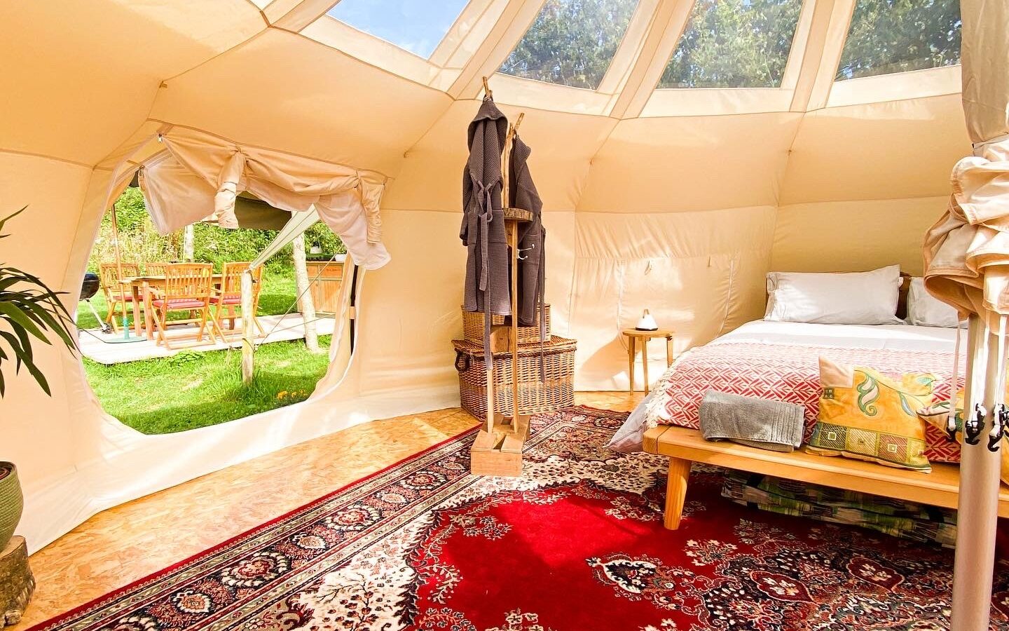 Interior of a luxury glamping tent on Coppet Hill featuring a cozy bed with colorful pillows and a blanket on a wooden frame. The tent has large clear panels letting in natural light. A Persian rug covers the floor, and in the background, an outdoor seating area is visible through the open tent flap.