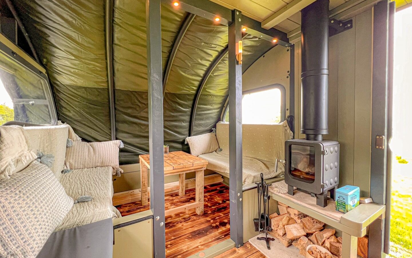 A cozy cabin interior on Coppet Hill, featuring a wood-burning stove with a small stack of firewood, flanked by two upholstered benches with pillows. A wooden table sits between the benches on a rustic wood floor. Warm lighting adds to the inviting atmosphere.