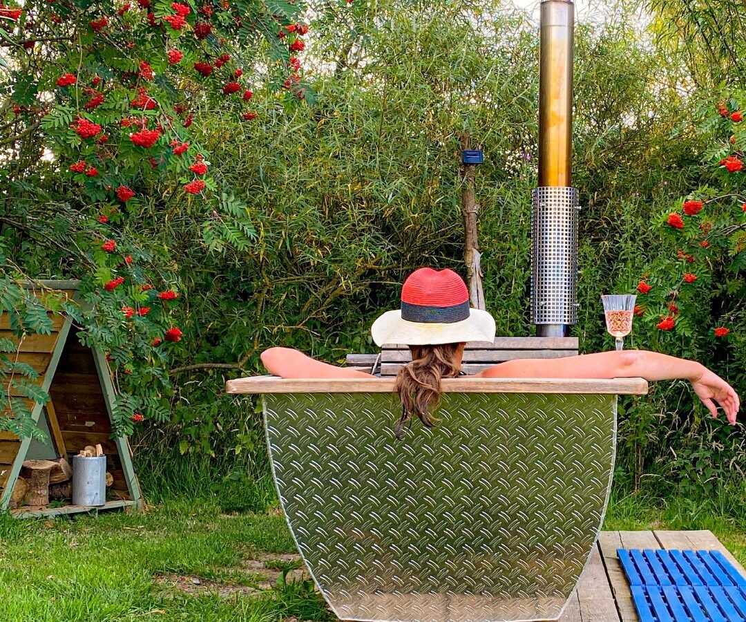 A person relaxes in an outdoor hot tub on Coppet Hill, surrounded by lush greenery and vibrant red flowers. Their arms rest on the sides of the tub, wearing a hat with a red band. A small table nearby holds a beverage, and a shed is visible in the background.