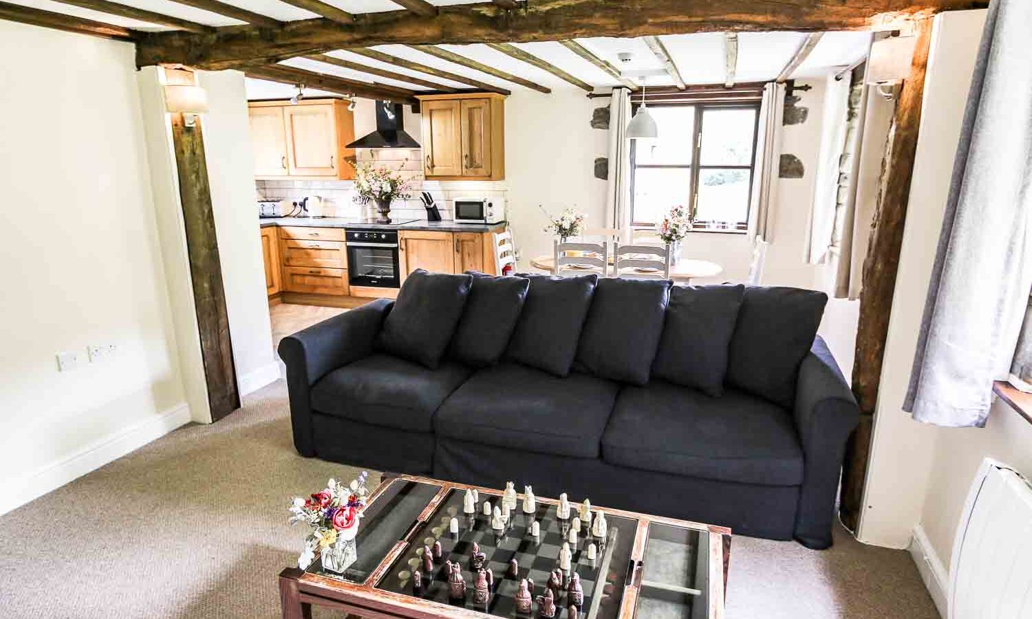 A cozy living room features a dark blue sofa with a glass coffee table in front of it, holding a chess set and small vases with flowers. The room opens into a kitchen with wooden cabinets, a stove, and a microwave. Exposed wooden beams and a large window add rustic charm, reminiscent of Cwm Chwefru cottages.