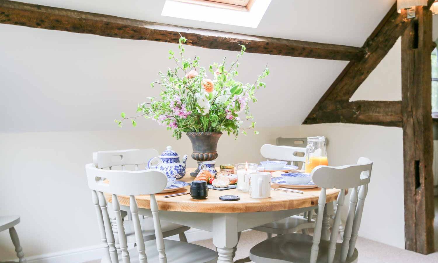 A cozy dining area in one of the charming Cwm Chwefru Cottages features a round wooden table surrounded by light gray chairs, adorned with a large bouquet of flowers in the center. The table is set with breakfast items, including juice, teapot, and baked goods. Exposed wooden beams and a skylight enhance the rustic charm.