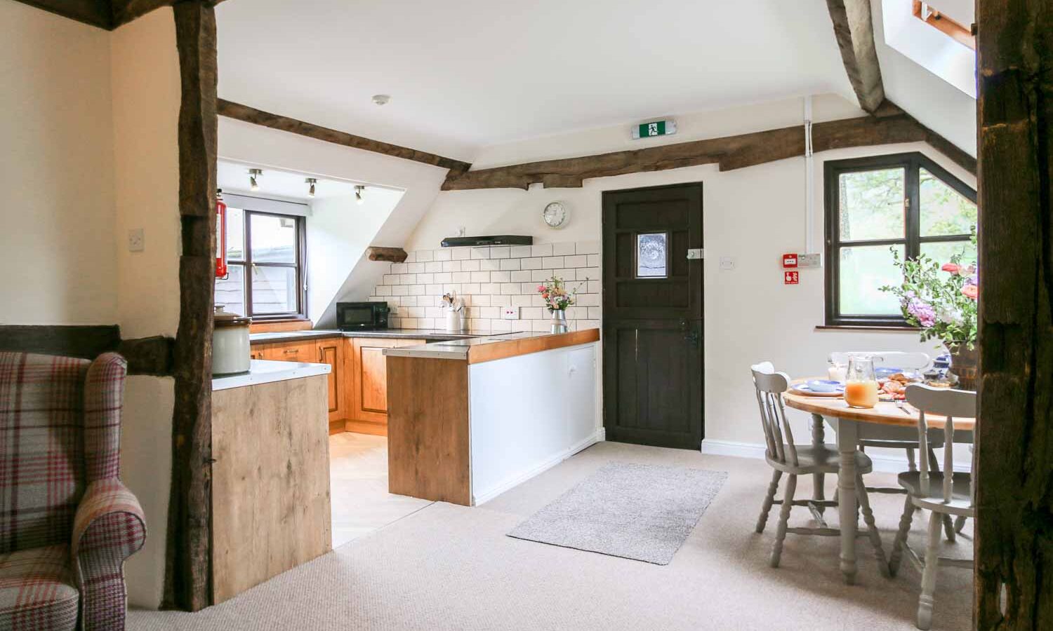 A cozy, rustic kitchen and dining area in the heart of Cwm Chwefru features wooden beams and white walls. The kitchen counter boasts wooden cabinets and modern appliances. A charming wooden dining table with chairs sits near a window adorned with flowers, creating a warm, inviting cottage atmosphere.