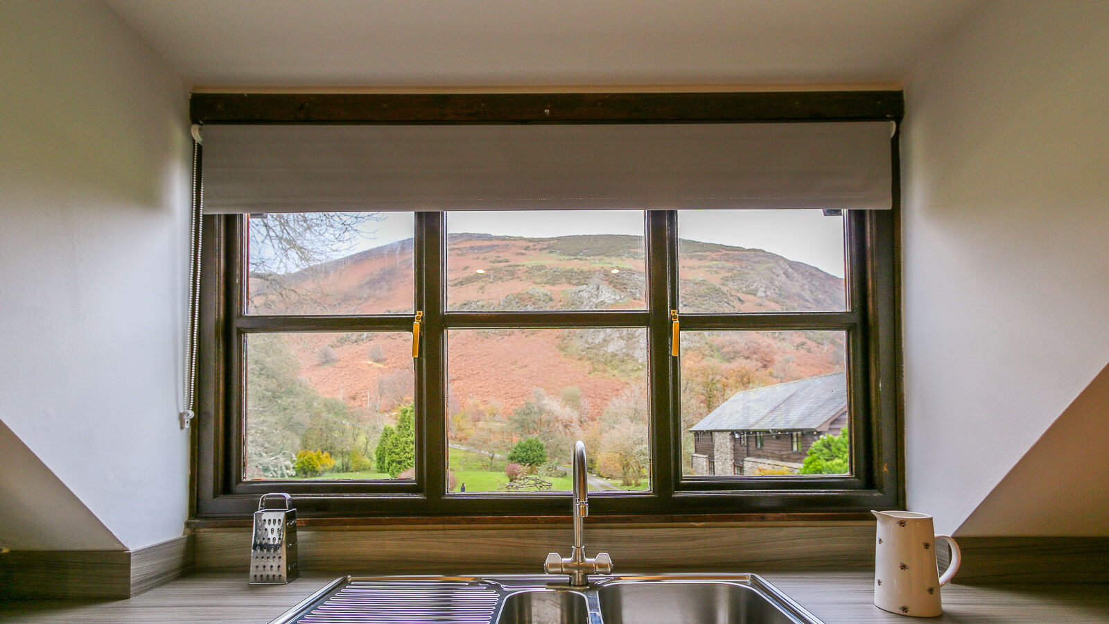 A kitchen window view of lush green fields at Cwm Chwefru Cottages, a house with a sloped roof, and a hillside with autumnal foliage. The countertop in the foreground has a stainless steel sink, a purple dish drying rack, a cheese grater, and a white ceramic pitcher.