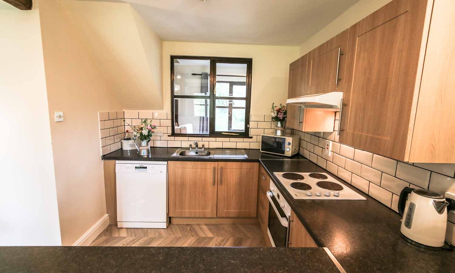 A modern kitchen with wooden cabinets, black countertops, and a beige tiled backsplash. It features an electric stove, dishwasher, microwave, and kettle. Two small windows above the sink overlook charming Cwm Chwefru Cottages, while decorative flower arrangements adorn the counter.