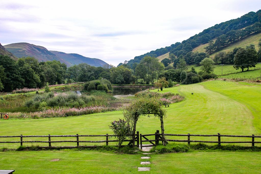 A lush green valley surrounded by gently sloping hills and dense trees features Cwm Chwefru cottages. The scene includes a well-maintained grass field, a small pond, and a wooden fence with a gate leading to a garden archway. The overcast sky adds to the serene and calm atmosphere.