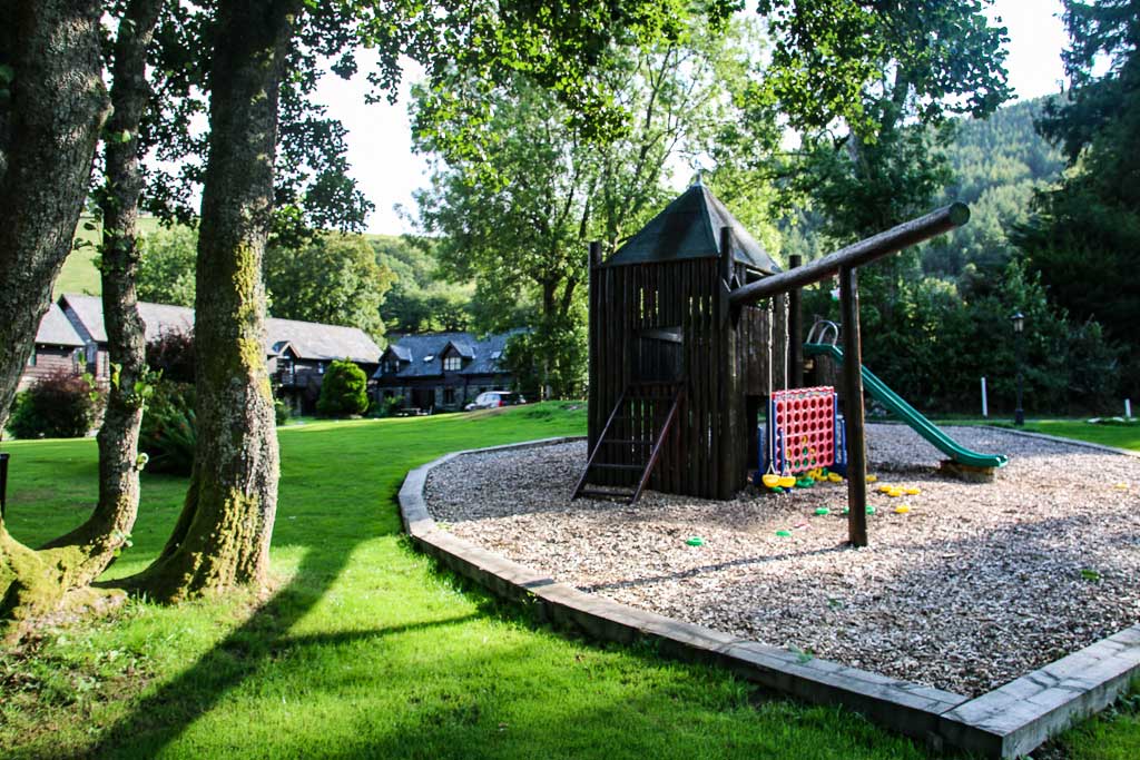 A wooden playground structure with a slide and climbing area stands on a bed of wood chips, surrounded by lush green grass and trees. In the background, there are charming rustic houses, reminiscent of Cwm Chwefru Cottages. Sunlight filters through the leaves, casting shadows on the ground.