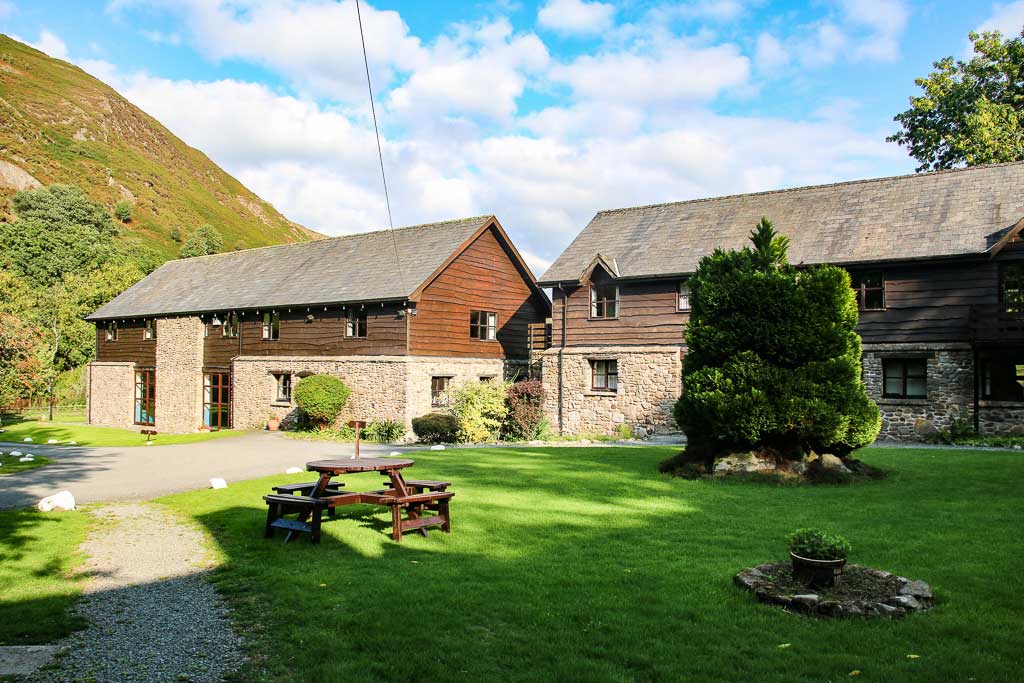 A picturesque scene of two rustic wooden and stone cottages set against a backdrop of green hills in Cwm Chwefru. The sky is partly cloudy. In the foreground is a well-manicured lawn with a picnic table and a neatly trimmed conifer tree.