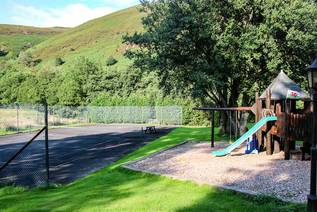 A scenic playground at Cwm Chwefru Cottages features a wooden play structure with a slide, set on a gravel base beside a lush green lawn. Adjacent to the playground is a fenced tennis court. The backdrop is a vibrant hillside covered with trees and dense foliage, under a clear sky.