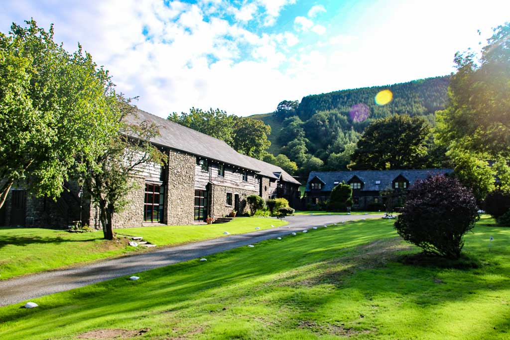 A tranquil scene of stone buildings with slate roofs nestled in lush greenery. A paved path winds through the well-manicured lawn, surrounded by trees and shrubs. Sunlight filters through the clouds, illuminating Cwm Chwefru Cottages against the scenic backdrop of hills and forests.