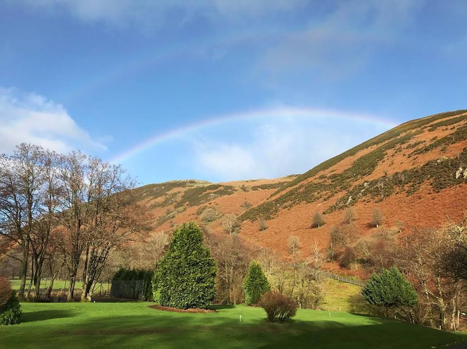 A scenic landscape featuring a green lawn in the foreground, flanked by clusters of trees on the sides. In the background, a hill covered with patches of brown vegetation rises under a blue sky with a faint rainbow arcing above it, reminiscent of the picturesque views near Cwm Chwefru Cottages.