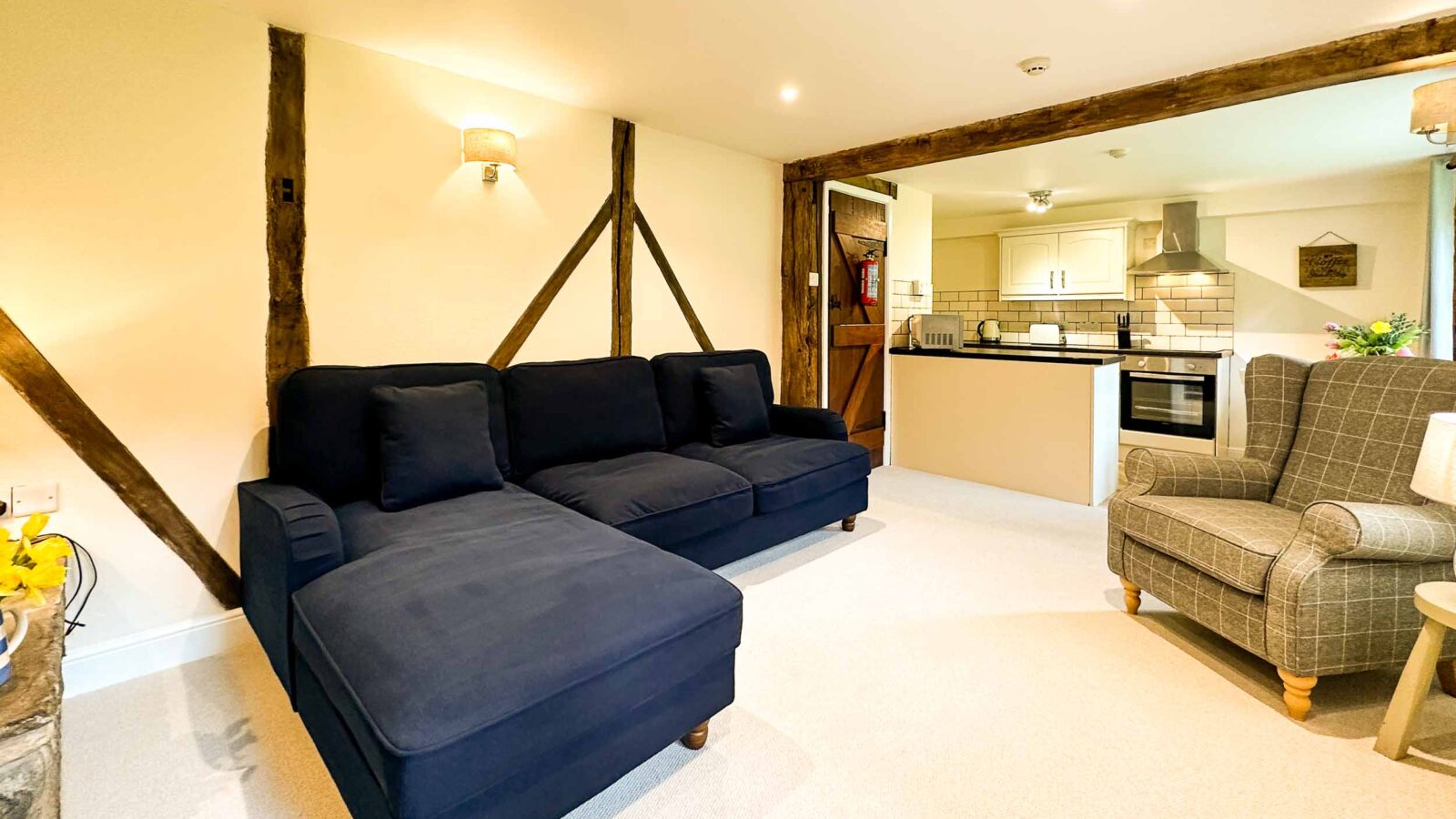 A cozy living room with exposed wooden beams features a large dark blue sectional sofa and a checkered armchair. In the background, there's an open-concept kitchen with light-colored cabinets, stainless steel appliances, and a small dining area, reminiscent of the charm found in Cwm Chwefru Cottages.