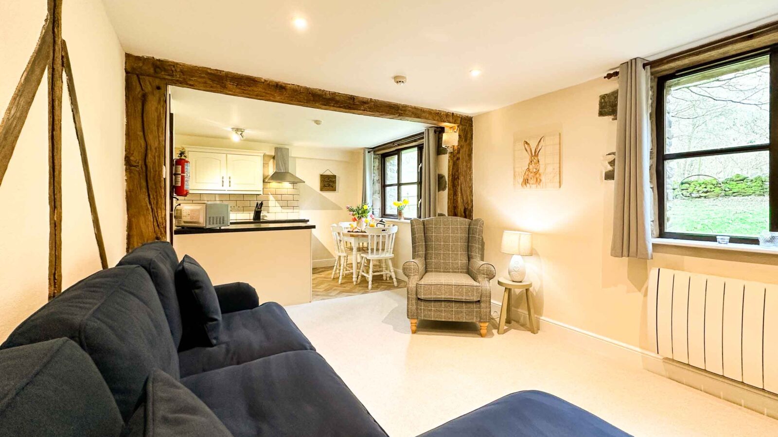 A cozy living area at Cwm Chwefru Cottages features a navy blue sofa, a gray armchair, and a small lamp in the corner. Wooden beams adorn the walls and a window with a curtain looks out to a green landscape. The space opens into a bright kitchen and dining area with a wooden table and white cabinets.