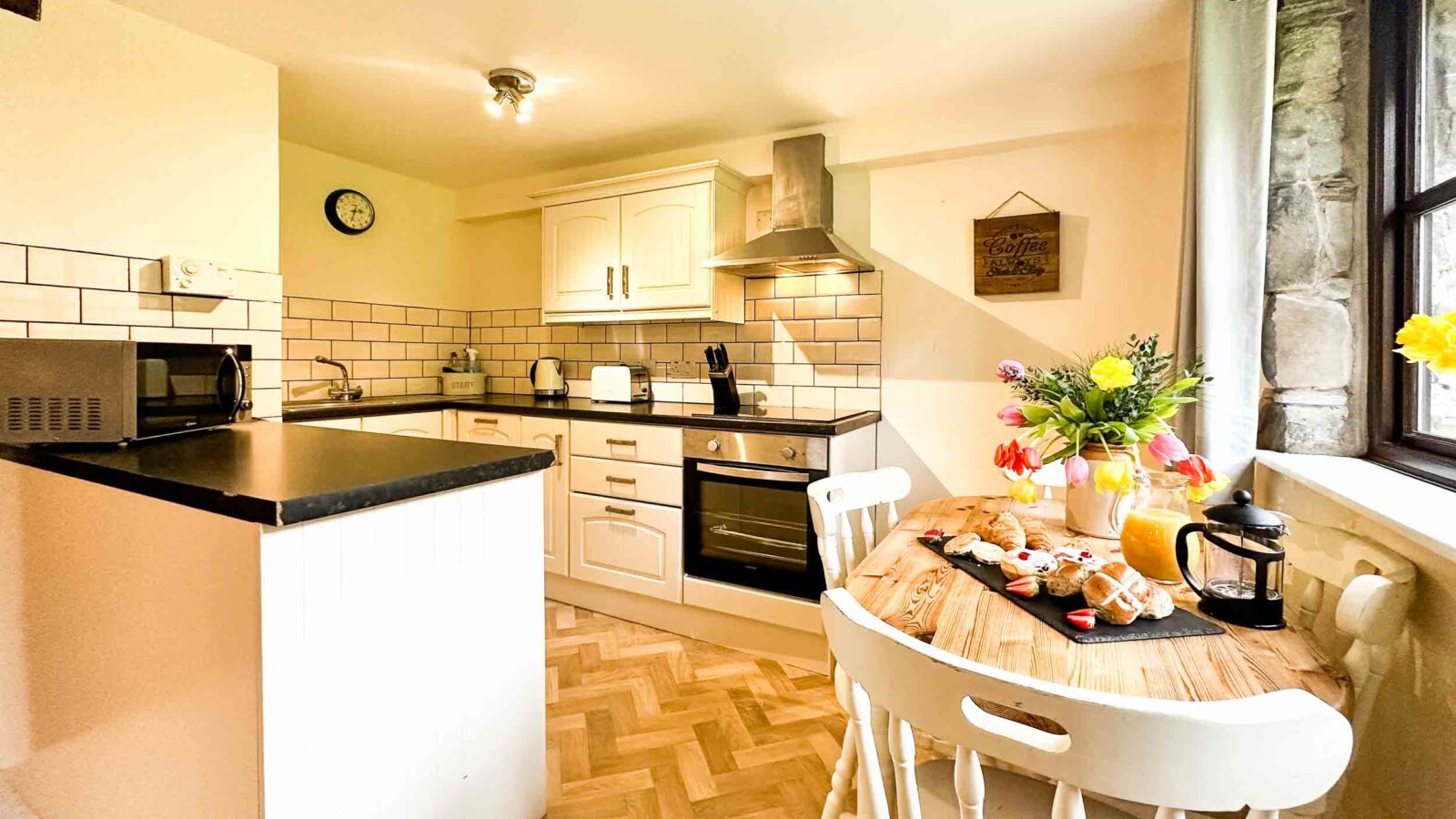 A cozy kitchen at Cwm Chwefru Cottages features wooden cabinets, a black countertop, and modern appliances, including a microwave and oven. A small round dining table with two chairs is set with coffee and pastries, adorned with a bouquet of flowers. A window brings in natural light.