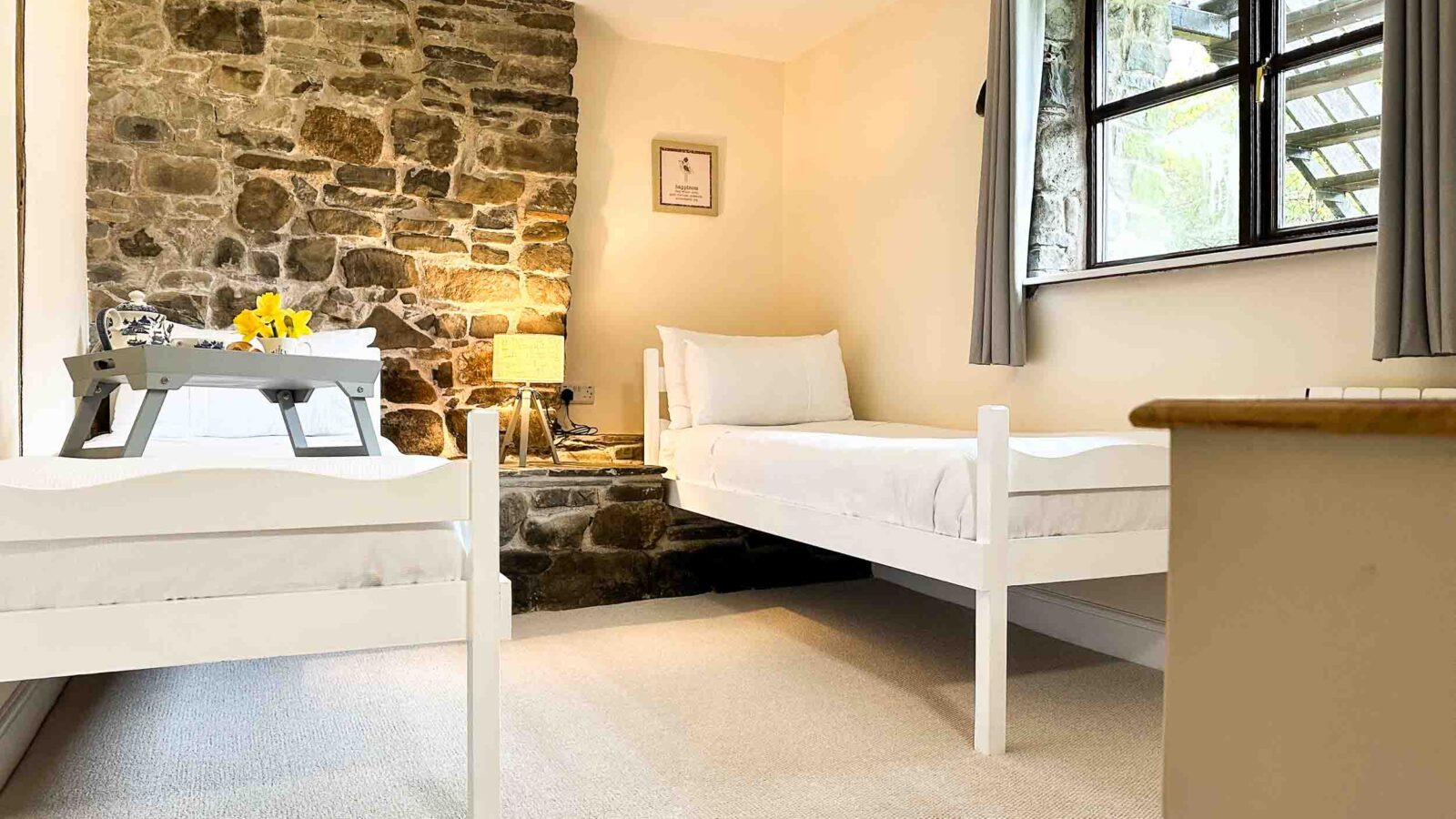 A cozy bedroom at Cwm Chwefru Cottages features two single beds with white frames and bedding, placed parallel to each other. There is a stone accent wall with a framed picture and a lamp on a wooden shelf. Sunlight streams through a window with gray curtains, creating a bright and inviting atmosphere.