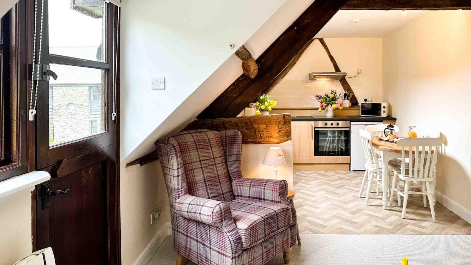 A cozy attic apartment in Cwm Chwefru features a plaid armchair beside a slanted wooden beam. The room includes a small dining table with white chairs, a modern kitchen with stainless steel appliances, and a vase of yellow flowers on the table, adding a cheerful touch to these charming cottages.