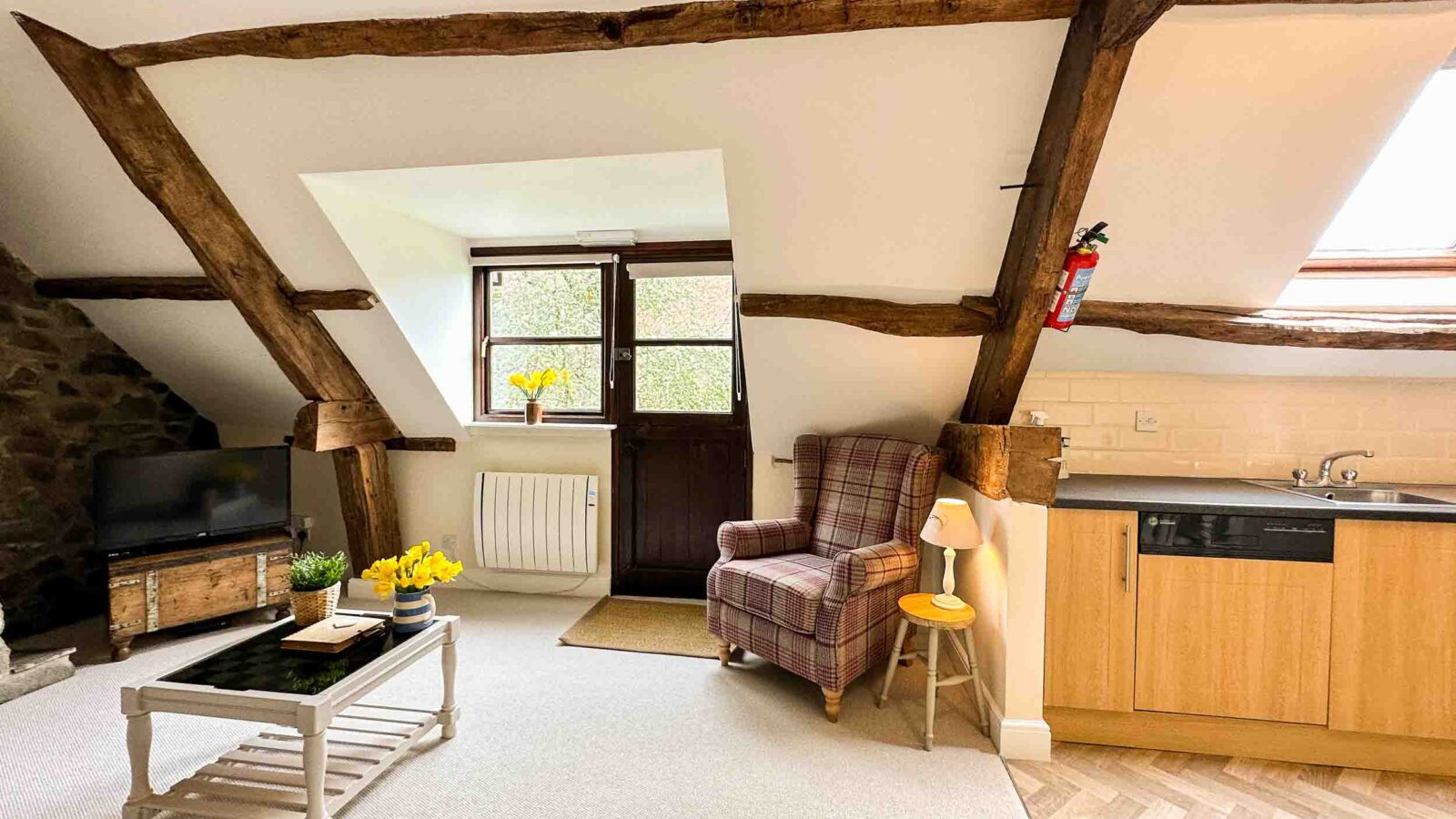 A cozy attic living space at Cwm Chwefru Cottages featuring exposed wooden beams, a slanted roof with a skylight, a small window, and a door. The room includes a plaid armchair, a TV, a white coffee table with a vase of daffodils, and a kitchenette with light wood cabinets.