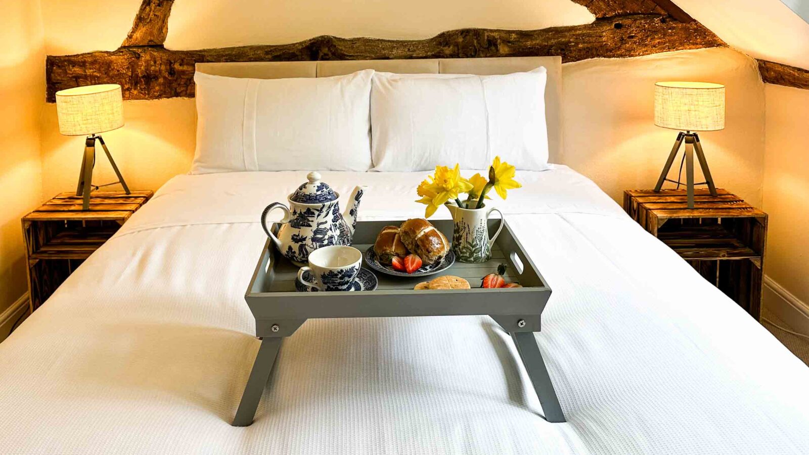 A cozy bedroom in Cwm Chwefru with a neatly made bed, adorned with a wooden tray holding a blue-and-white teapot, cup, saucer, croissants, a small bowl of spreads, and a vase of daffodils. Rustic wooden beams decorate the wall, and two lit bedside lamps sit on wooden nightstands, capturing the charm of cottages.