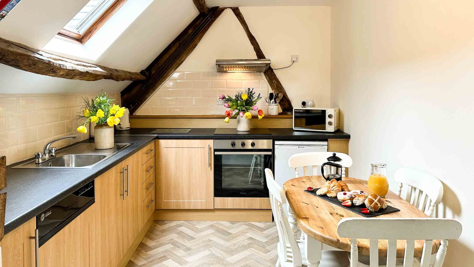 A cozy kitchen at Cwm Chwefru Cottages boasts light wood cabinets, a black countertop, and a skylight. It features an electric stove, oven, microwave, and sink. A round wooden table with white chairs is set for breakfast with orange juice, fruits, and pastries. Bright flowers add a decorative touch.