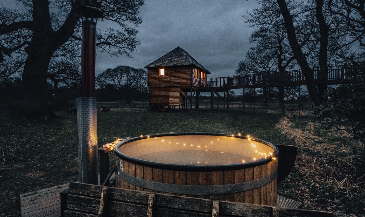 A wooden hot tub is illuminated with string lights against a backdrop of a dark, wooded area. In the distance, the Netherby Treehouse, a charming two-story cabin elevated on stilts and adorned with lights, adds to the evening's magic under an overcast sky and silhouetted trees.