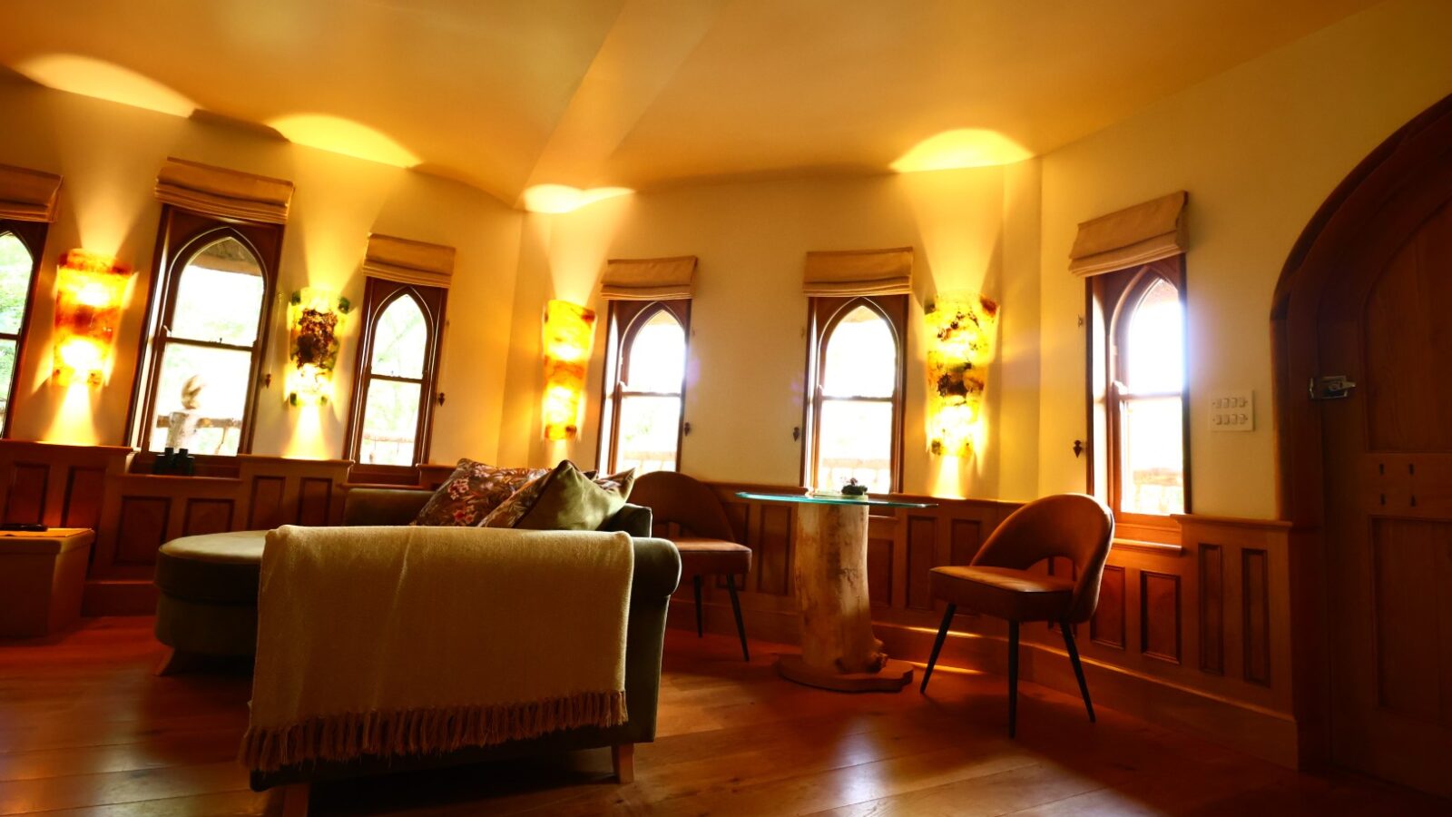 A cozy Treeopia-themed room with arched windows adorned with elegant shades and wall-mounted lights. The room features wooden flooring, a comfortable curved sofa draped with a blanket, a round table, and two chairs. The warm lighting creates an inviting ambiance.