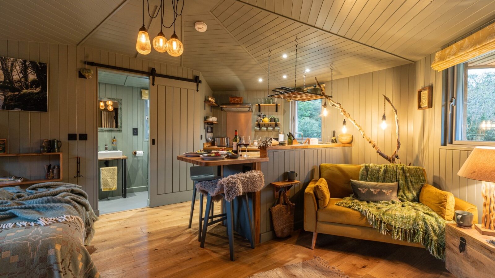 The Cabin, an inviting haven by Erwain Escapes, boasts a cozy, well-lit interior with wooden panels and floors. It features a yellow sofa accented by green throw blankets, a bed with patterned blankets, a small kitchen with stools, and rustic decor including hanging lights, wooden shelves, plants, and artwork.