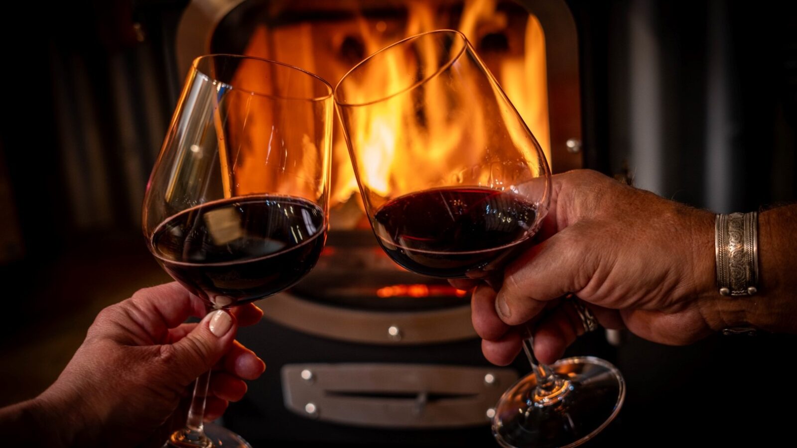 Two hands holding glasses of red wine engage in a toast in front of a roaring fireplace at Erwain Escapes. The warm glow of the fire reflects off the wine, creating an inviting and cozy atmosphere reminiscent of a night spent in The Cabin.