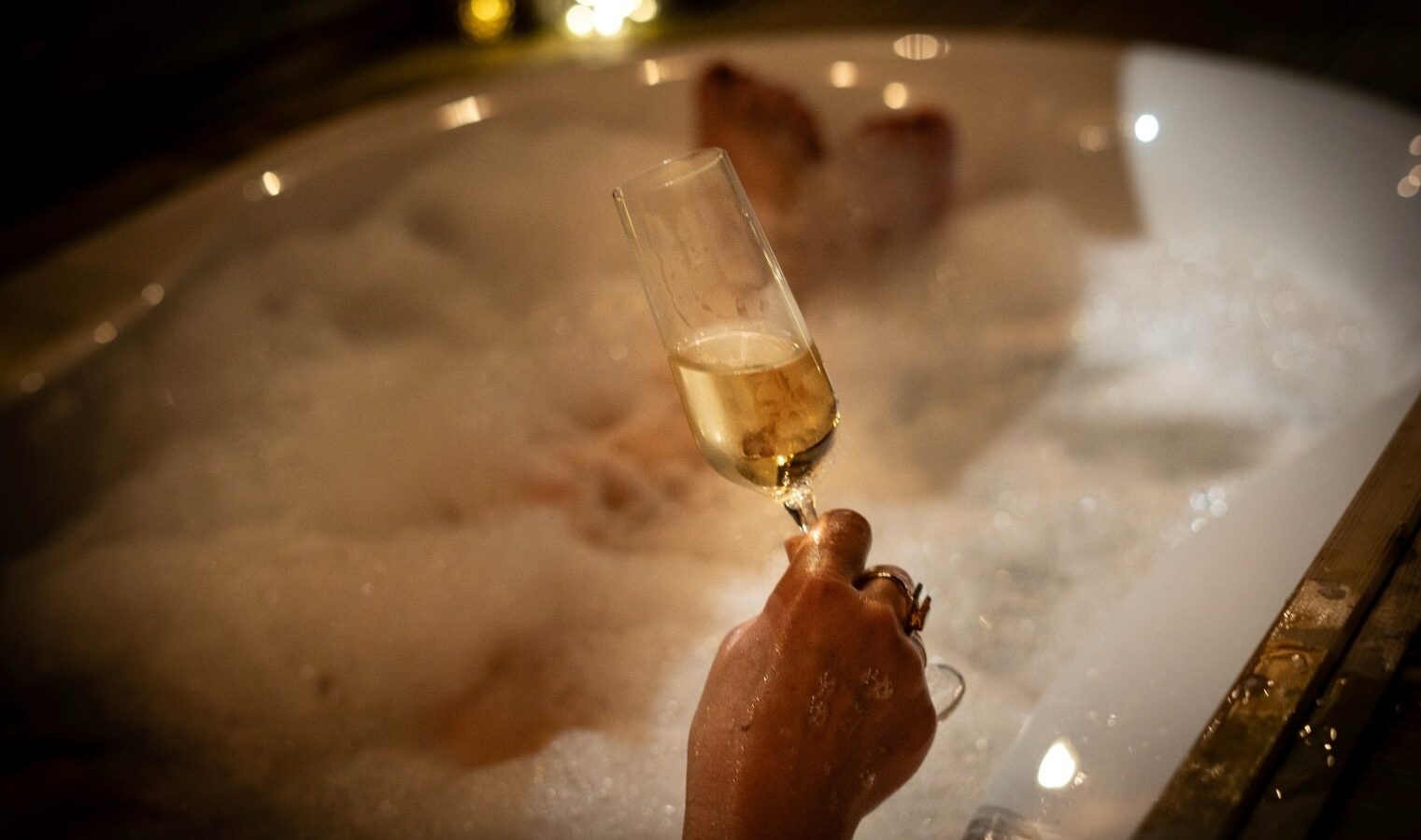 A person relaxes in a bubble-filled bathtub at Erwain Escapes, holding a glass of champagne. The soft lighting and bubbles create a calming and luxurious atmosphere, perfect for unwinding after exploring The Cabin's enchanting surroundings.