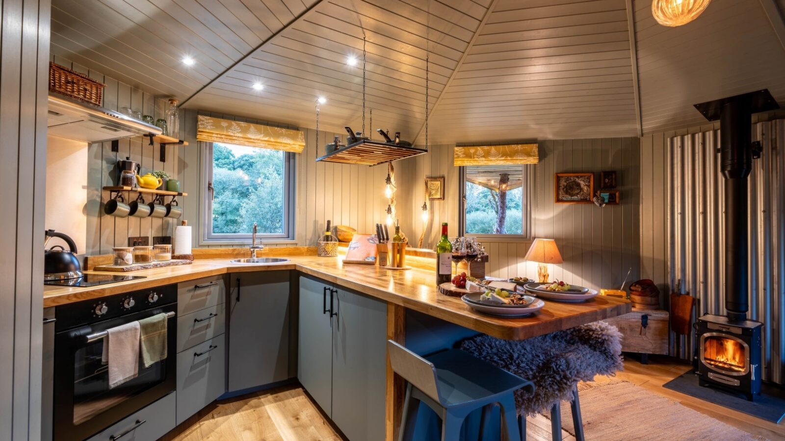 Cozy kitchen interior with wood-paneled walls and ceiling, featuring a stove, oven, and wooden countertops. A rustic dining area with a table set for two is seen next to a window. A lit stove and warm lighting create a homely atmosphere in the corner—a perfect retreat from Erwain Escapes at The Cabin.