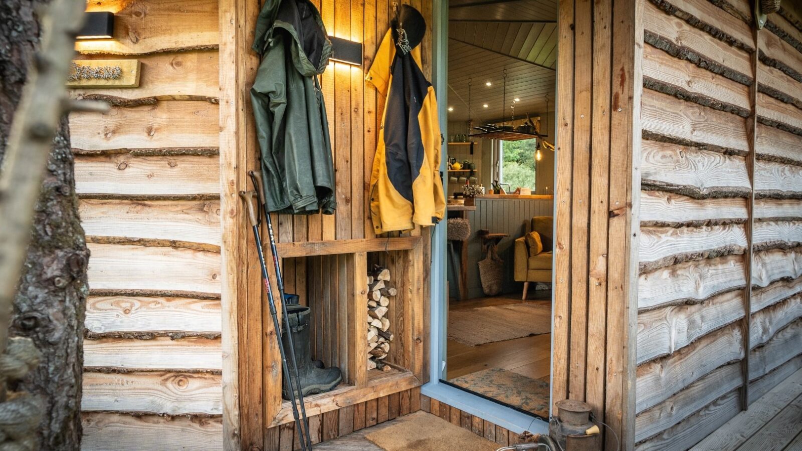 A cozy wooden cabin entrance at Erwain Escapes greets you with hanging raincoats, walking sticks, and boots. Through the open door of The Cabin, a warmly lit interior with a comfortable seating area is visible. Firewood is neatly stacked in an alcove beside the door. Outside, a basket of logs and kindling sits on the wooden porch.