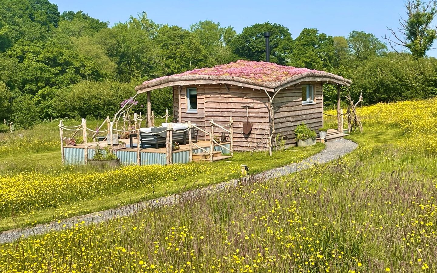 A small, round wooden cabin with a green roof stands in a lush meadow filled with yellow wildflowers. A winding gravel path leads to the Erwain Escapes retreat, which boasts a fenced wooden deck area. The background features dense, green trees under a clear blue sky.