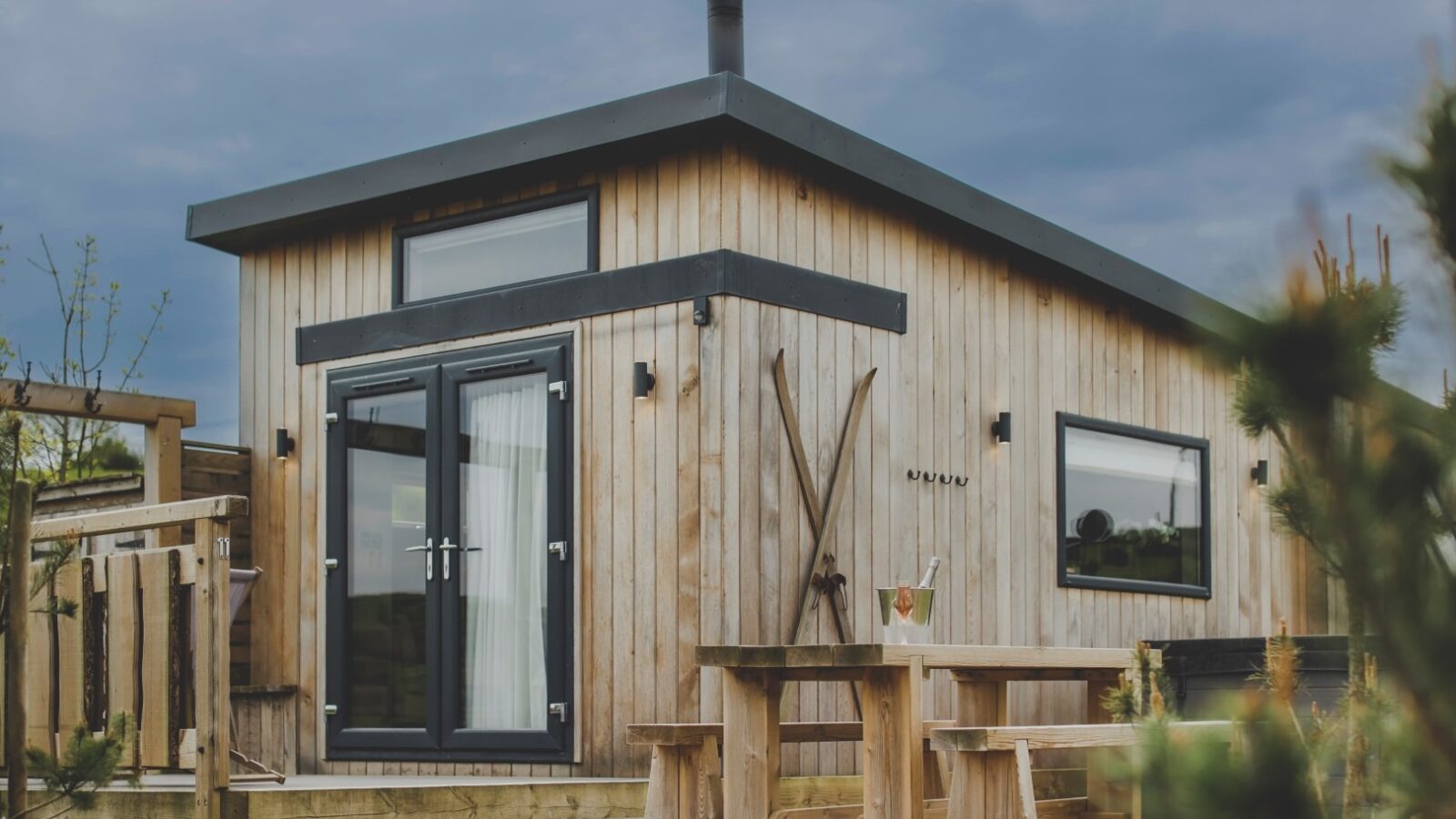 A modern, small wooden tiny house with large windows and a slanted roof stands as a perfect retreat. In front of the house is a rustic wooden table with benches and potted plants around it. Skis lean against the exterior wall, and the sky is cloudy in the background.