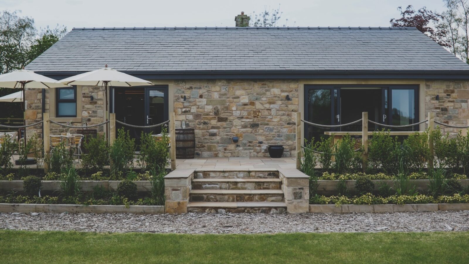 A quaint stone cottage with a grey slate roof sits behind a meticulously landscaped garden. This Everything Retreat features large windows and a central entrance with steps leading up, flanked by rope handrails. Two patio umbrellas are visible on the left side.