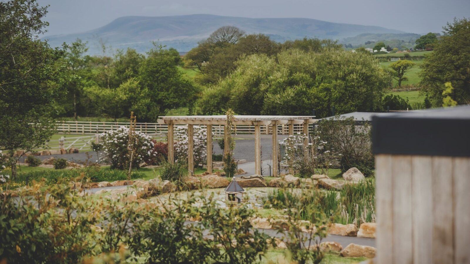 A lush garden retreat features a wooden pergola surrounded by greenery and a small pond. In the background, a mountainous landscape stretches under a cloudy sky. Fences and scattered trees dot the landscape, adding to the serene and picturesque setting of this everything retreat.