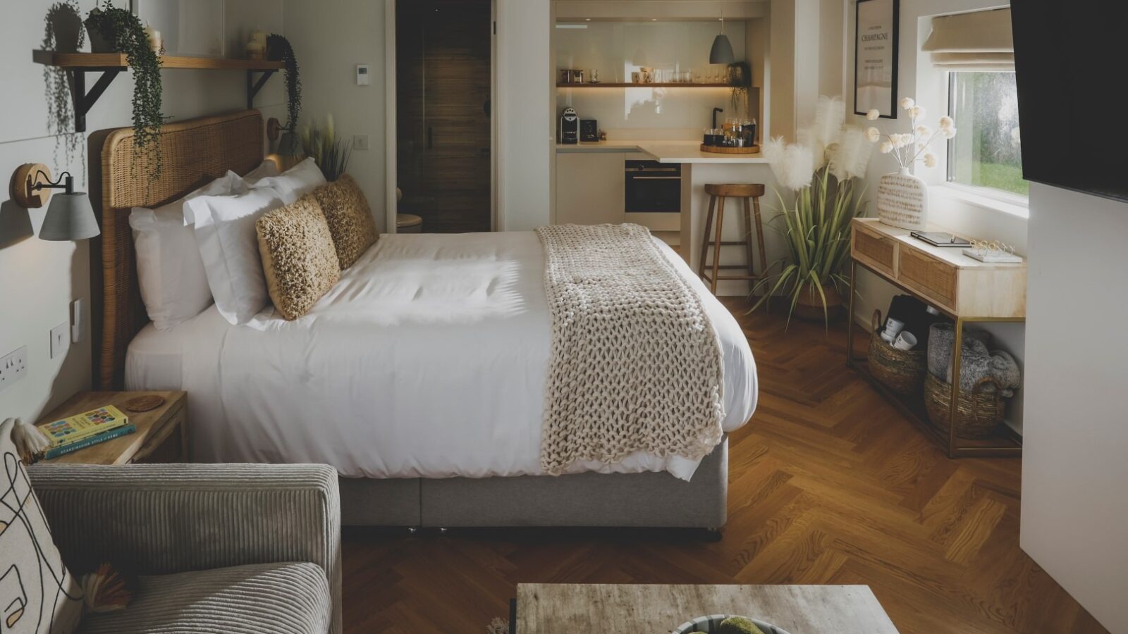 A cozy, modern bedroom retreat with a neatly made bed featuring beige and white bedding. The room has wooden floors, a small kitchen area in the corner, a sofa, a wooden desk near the window, and various plants and decorative items adding a warm touch to everything.