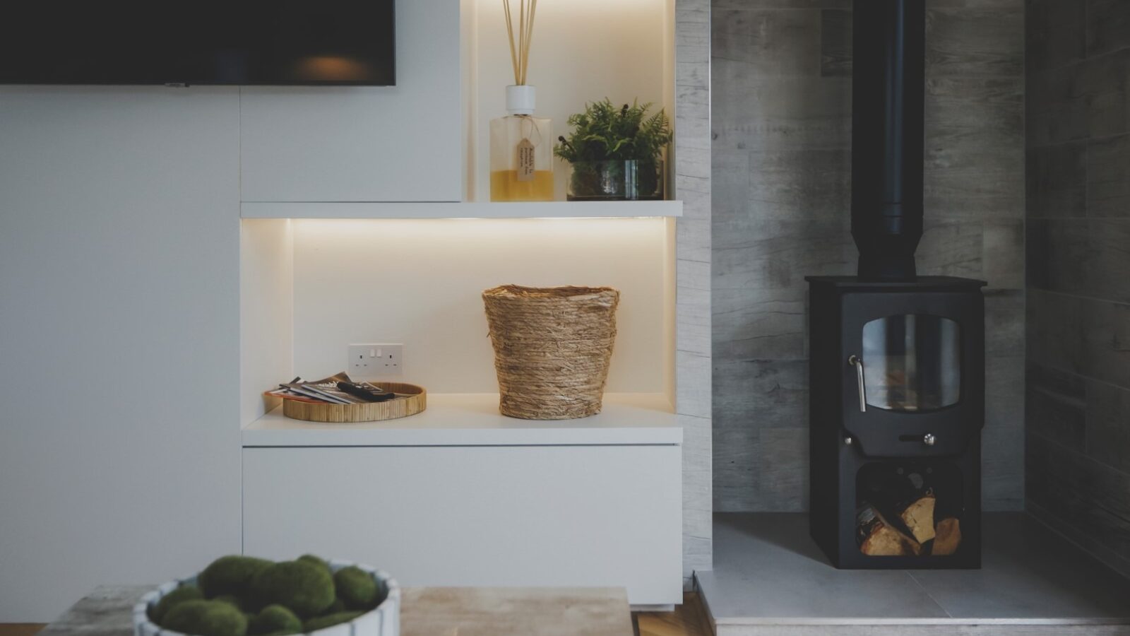 A modern living room corner serving as the perfect retreat, featuring a black wood-burning stove with a vertical glass door on the right. On the left, built-in white shelves hold a wicker basket, plants, and decorative items. A bowl of green decorative spheres is visible in the foreground.