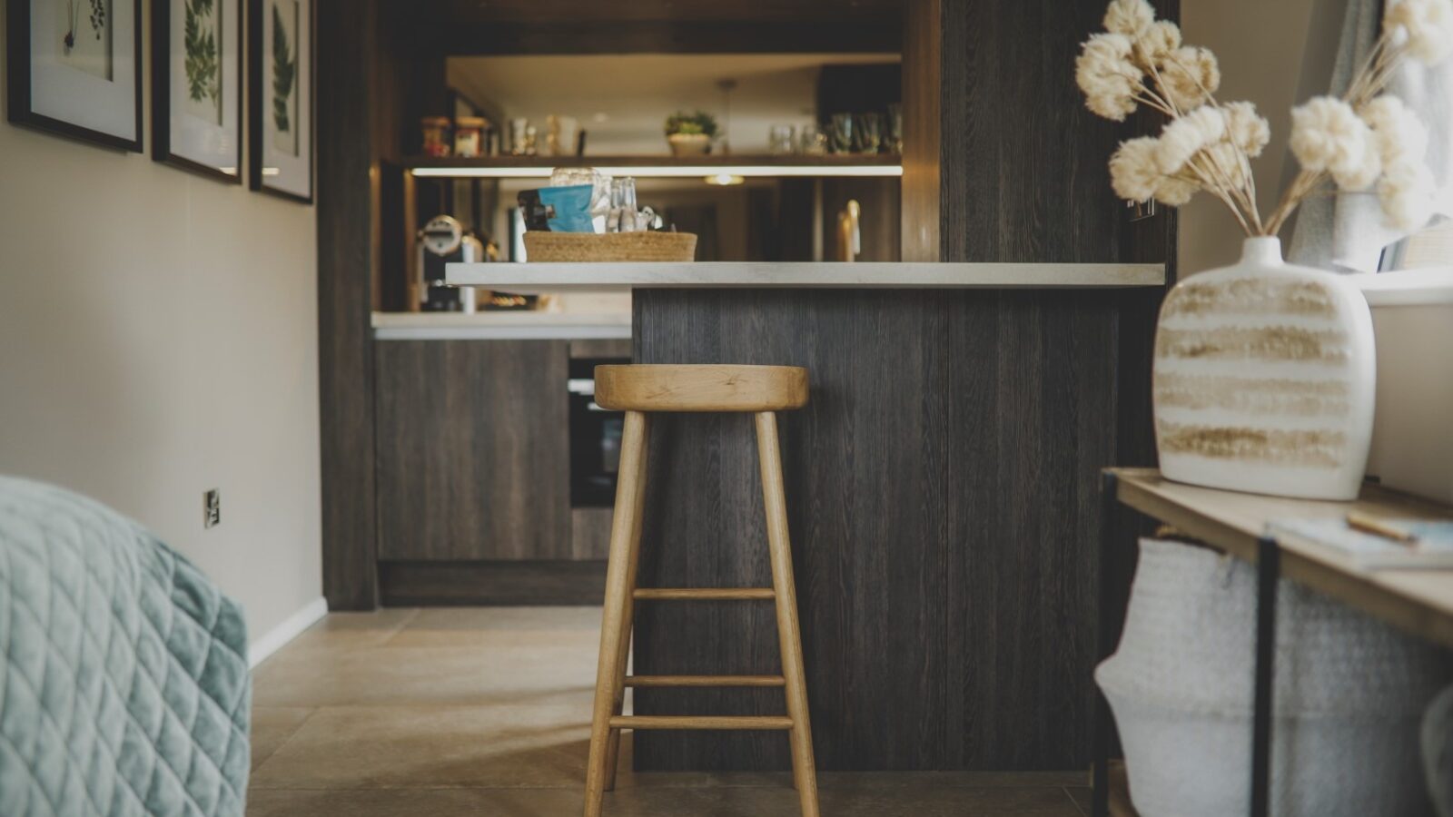 A modern kitchen with dark wood cabinets and a white countertop. A wooden bar stool is placed near the counter, making it an ideal spot for a relaxing retreat. The room is decorated with framed botanical prints on the wall, a vase with dried flowers, and a light fixture hanging from the ceiling.