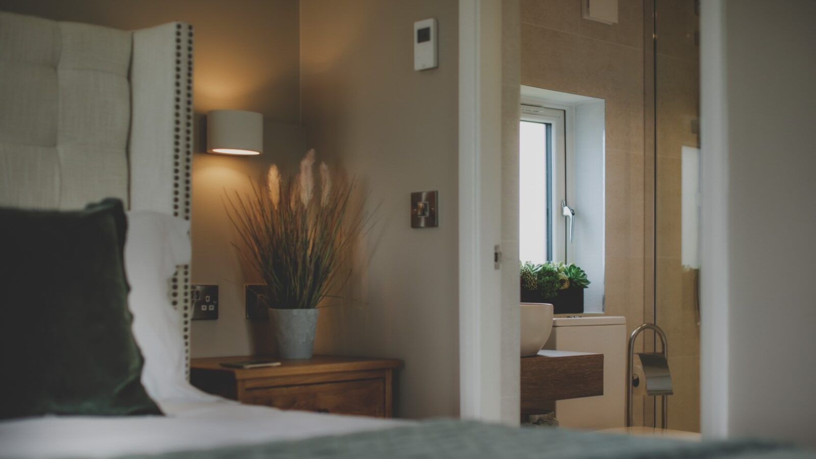 A cozy bedroom with a bed featuring a dark green pillow, a bedside table with a potted plant, and a neutral-colored wall. An open door reveals a bathroom with a white sink, some greenery on the windowsill, and a glass-enclosed shower. Perfect for an Everything Retreat.