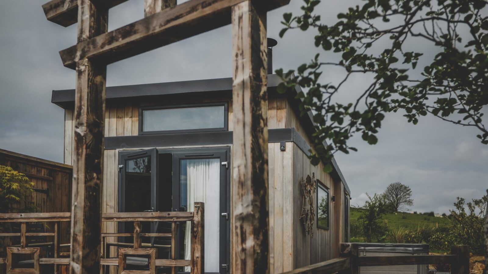 A small, modern wooden cabin with large windows and a glass door, framed by a rustic wooden archway. This serene retreat is surrounded by greenery and trees, with a cloudy sky in the background.