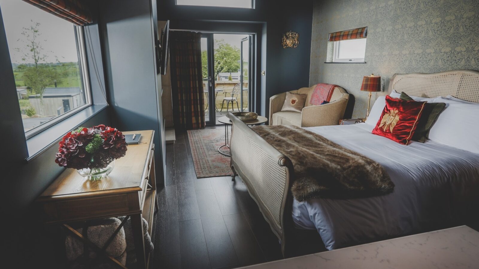 A cozy bedroom with dark walls features a large bed adorned with a plush fur throw and vibrant pillows, epitomizing the essence of an Everything Retreat. Beige armchairs sit beside a small table, while natural light floods in through the window and glass door, revealing views of the green landscape. A vase with red flowers graces the wooden desk.