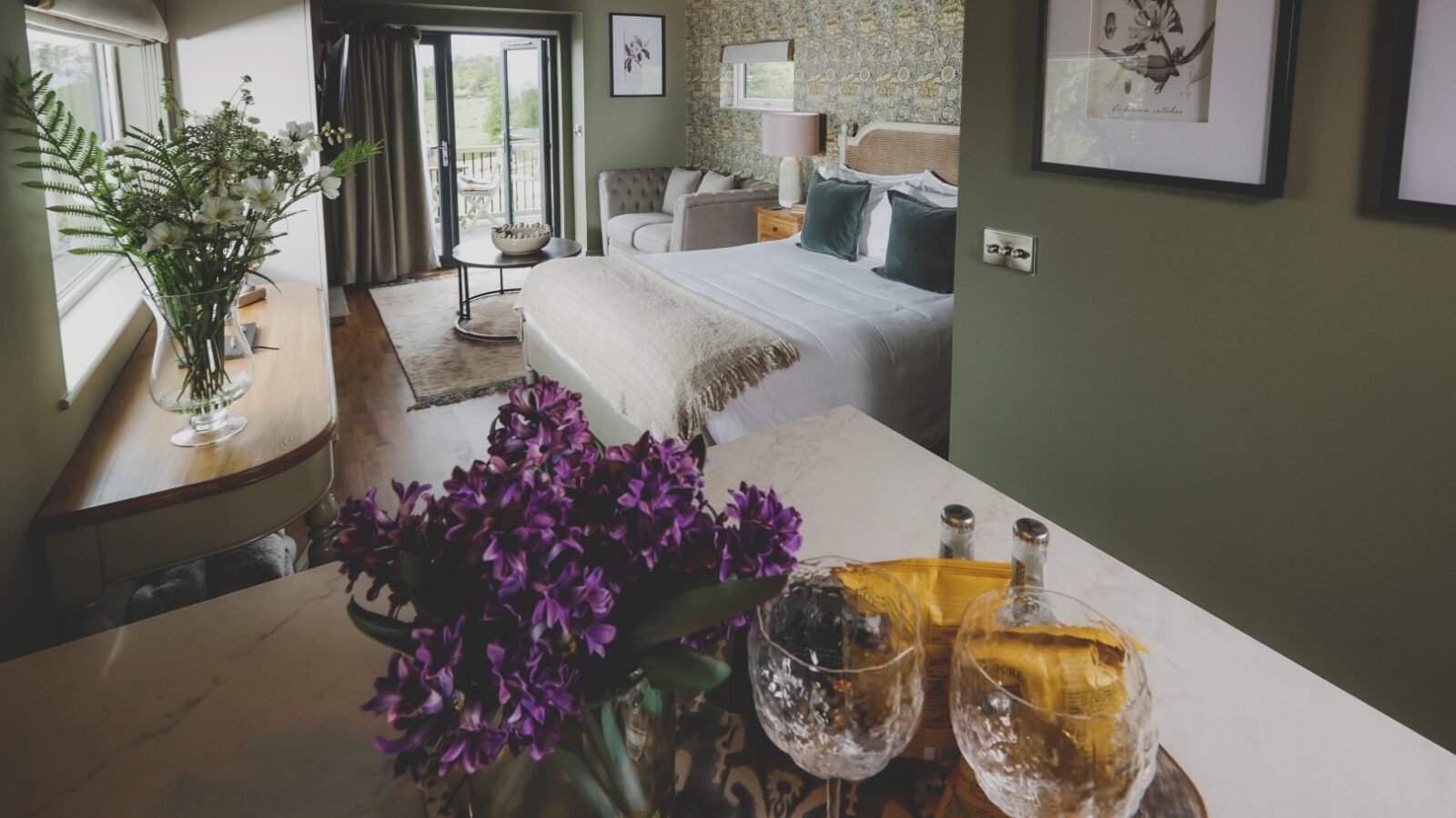 A beautifully designed bedroom with a green color palette features a neatly made bed with a beige headboard. At this Everything Retreat, there's a cozy seating area by a large window with a garden view. In the foreground, purple flowers and two crystal glasses sit on a tray on the table.