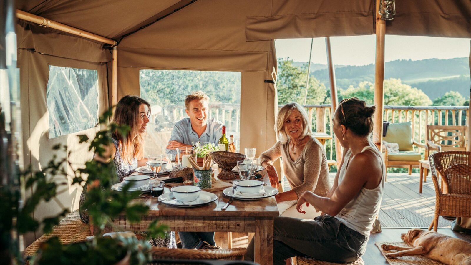 A group of four people enjoys a meal together inside a cozy, tented outdoor dining area at Exe Valley Glamping. The sun shines through the open sides, illuminating their smiles as they converse around a rustic wooden table set with plates, glasses, and a wine bottle.