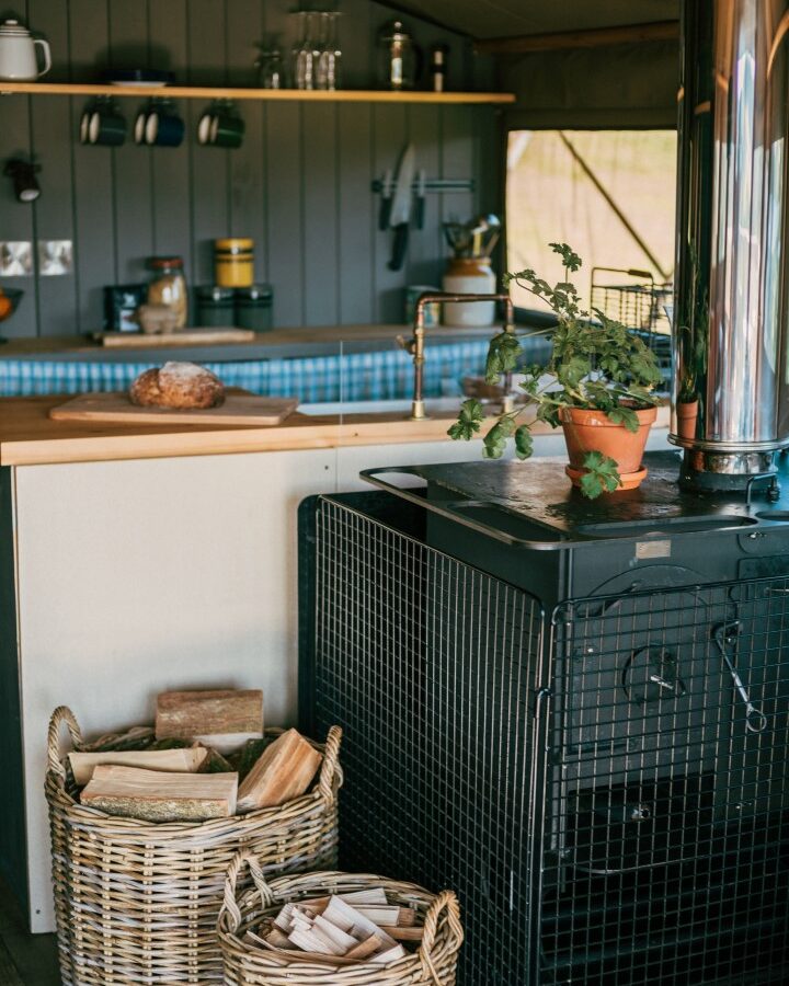 A cozy kitchen at Exe Valley Glamping features a wood-burning stove. Two wicker baskets filled with firewood sit on the floor. A potted plant is on the stove, surrounded by wooden shelves with jars and cups, a loaf of bread on a cutting board, and a checkered blue backsplash.