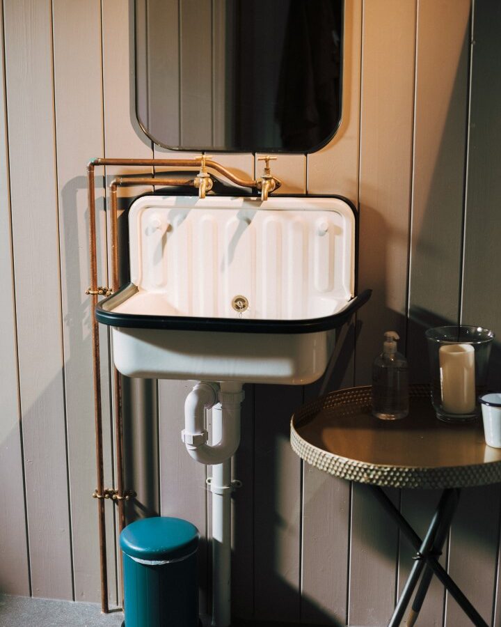 A small, vintage-style bathroom sink with exposed bronze plumbing is mounted on a wooden wall at the cozy Exe Valley glamping site. Above the sink is a mirror. To the right, a gold-toned round table holds a soap dispenser, a candle, and a small cup. A teal-colored trash bin is under the sink.