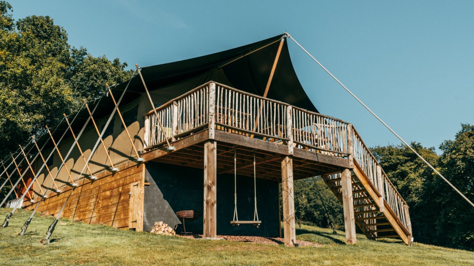 A modern, elevated tent structure with a wooden deck and stairs sits in the green grassy expanse of Exe Valley, surrounded by trees. Ideal for glamping, the setup includes a swing hanging from the platform and a barbecue grill beneath the deck. The sky is clear and blue.