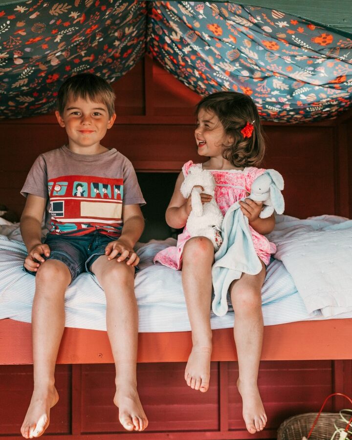 Two young children sit on a bed. The boy on the left wears a t-shirt and shorts, while the girl on the right, dressed in a pink dress, laughs while holding stuffed animals. They sit in a cozy, playhouse-like space with a colorful, patterned fabric ceiling—ideal for Glamping in Exe Valley.