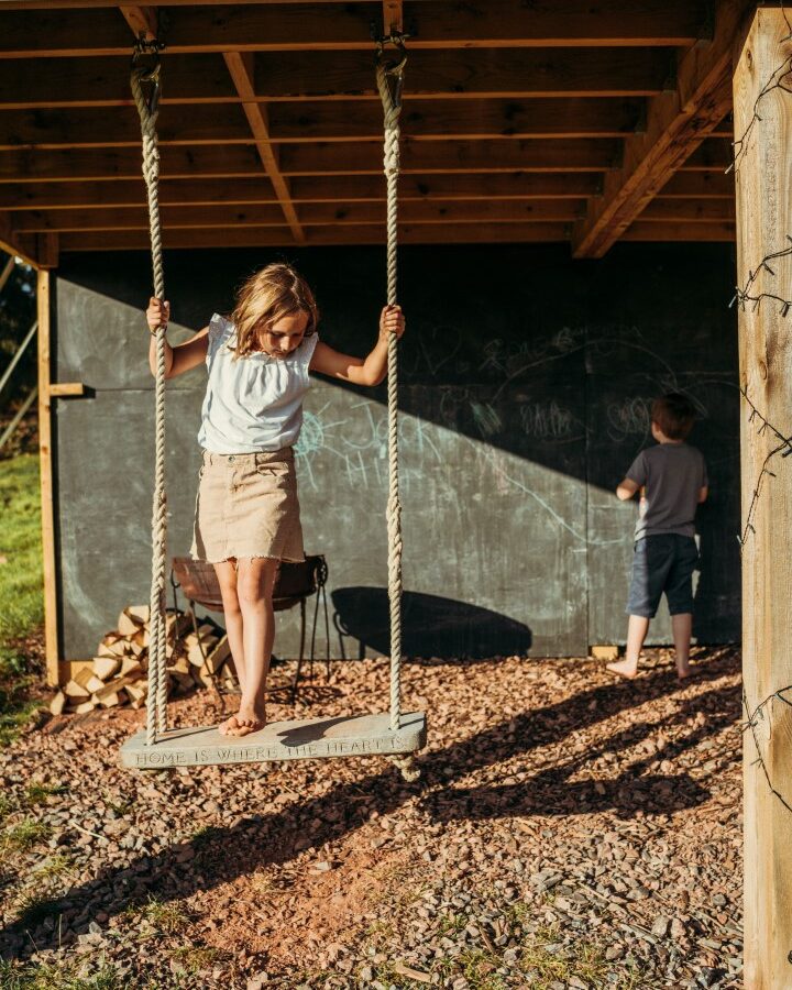 A young girl stands on a swing with wooden planks, hanging from ropes, under a wooden structure at Exe Valley Glamping. She is focused on maintaining her balance. In the background, a boy writes or draws on a large chalkboard attached to the wall. Logs are stacked to the side.