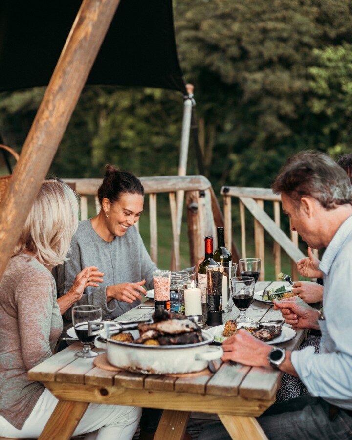 A group of four people sits around a wooden picnic table outdoors at Exe Valley Glamping, enjoying a meal together. They are engaged in conversation and laughing. Various dishes, wine glasses, and bottles are on the table. The atmosphere appears relaxed and cheerful, with trees visible in the background.