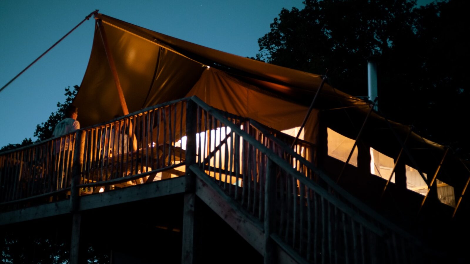 A large tan tent is set up on an elevated wooden platform at dusk, offering the perfect Exe Valley Glamping experience. Lights from inside the tent cast a warm glow. A person stands near the edge of the platform, partially silhouetted, with trees and a twilight sky in the background.