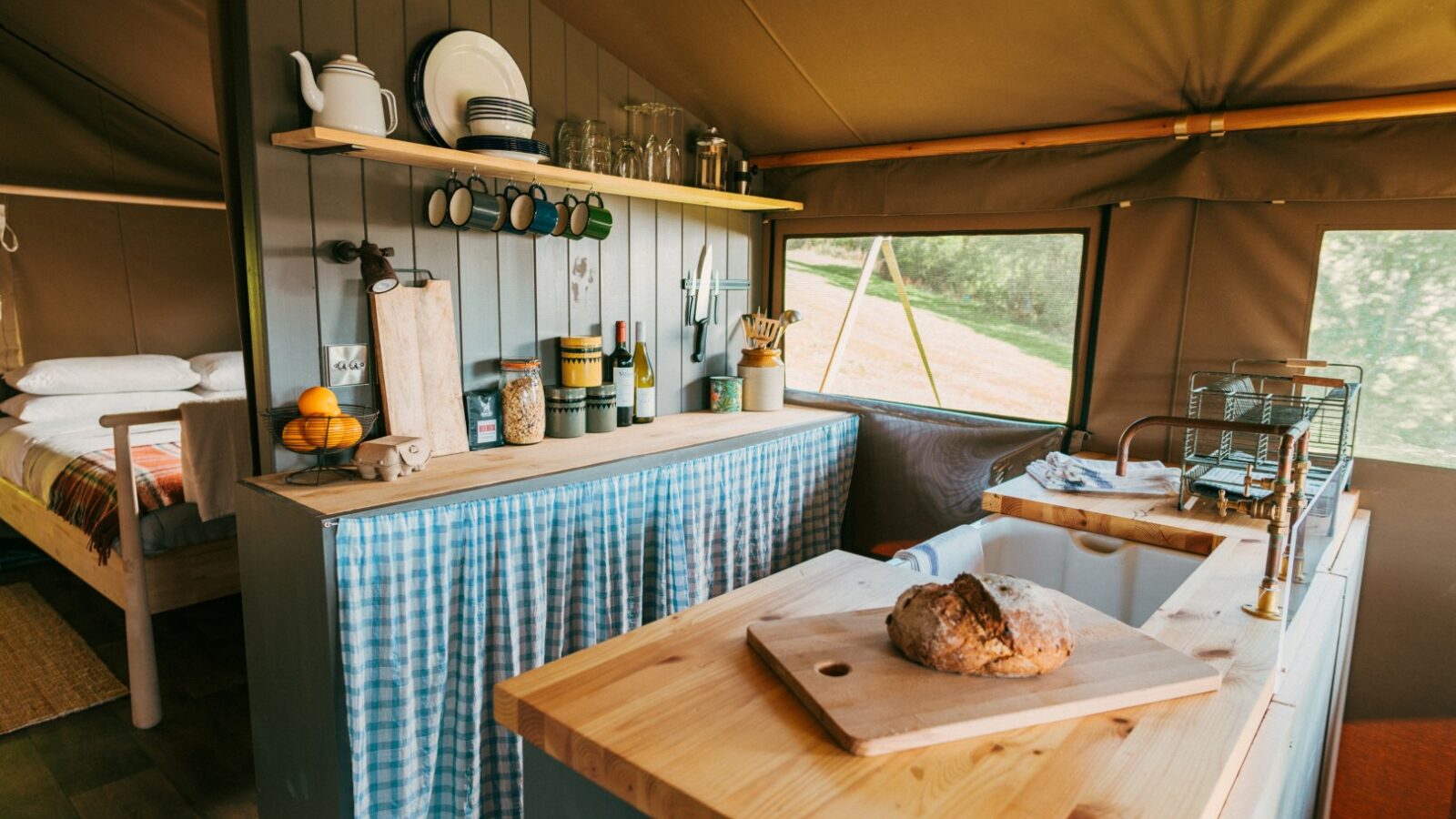 A cozy glamping kitchen with a wooden countertop, featuring a loaf of bread on a cutting board and various kitchen items. Shelves with cups and dishes are above the counter, revealing an exquisite green outdoor view of the Exe Valley through the window. A bed is partially visible in the background.