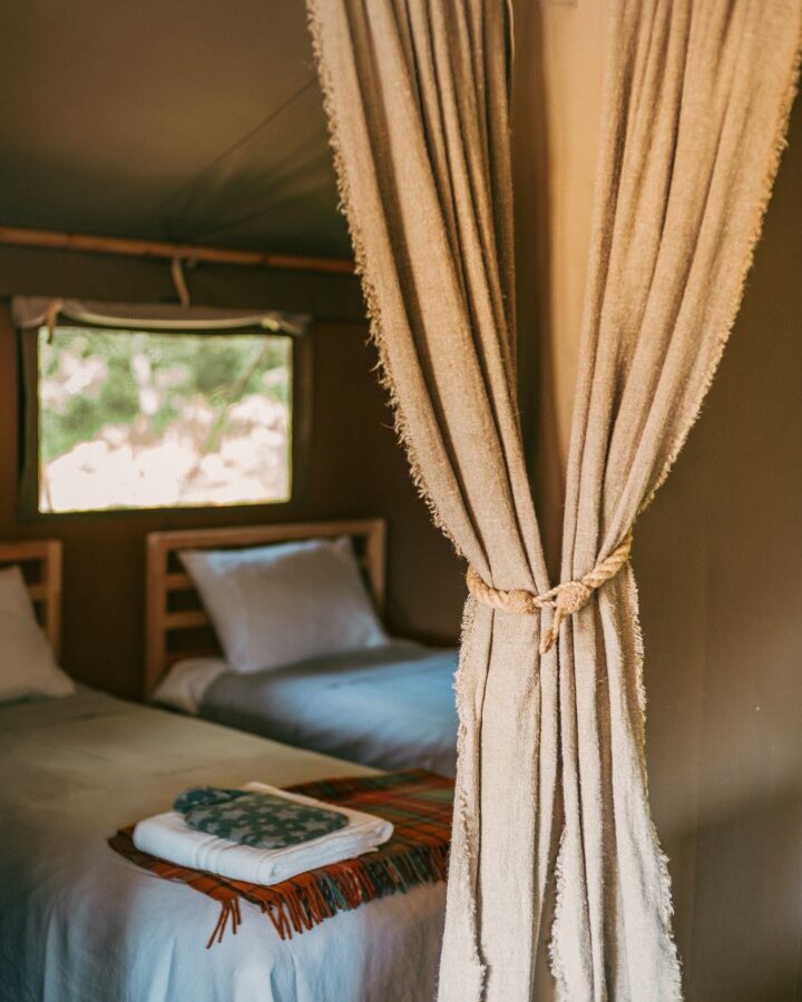 A cozy tent interior at Exe Valley Glamping features two neatly made beds with white linens, separated by a tied-back beige curtain. A small window above one bed lets in natural light, and both beds have folded blankets and towels placed on them, adding a touch of comfort.